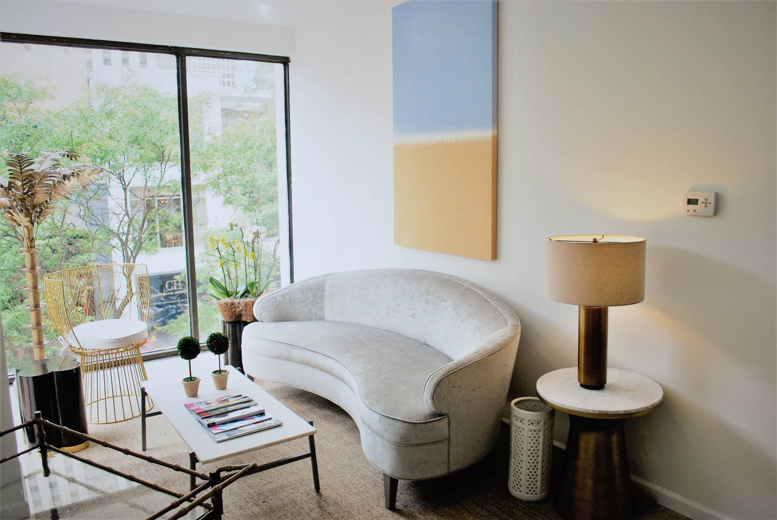 A modern living room with a curved gray sofa, round side table with lamp, glass coffee table, and large window overlooking greenery.