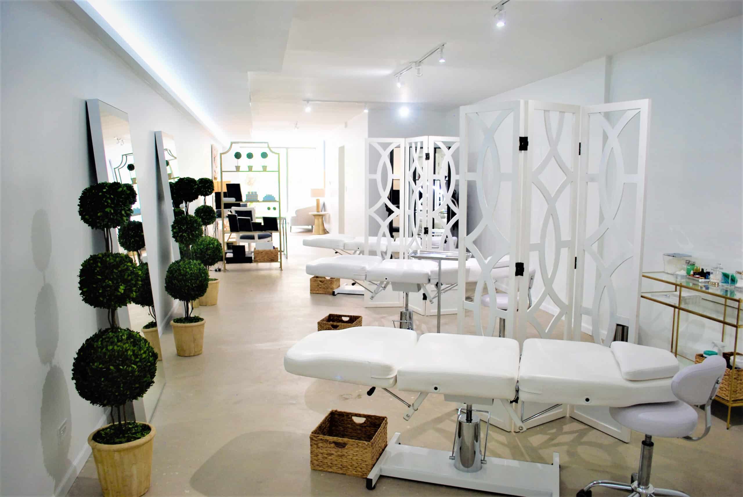 A modern spa room with white treatment beds, privacy screens, potted topiary plants, mirrors, and wicker baskets, all in a clean, minimalist setting.