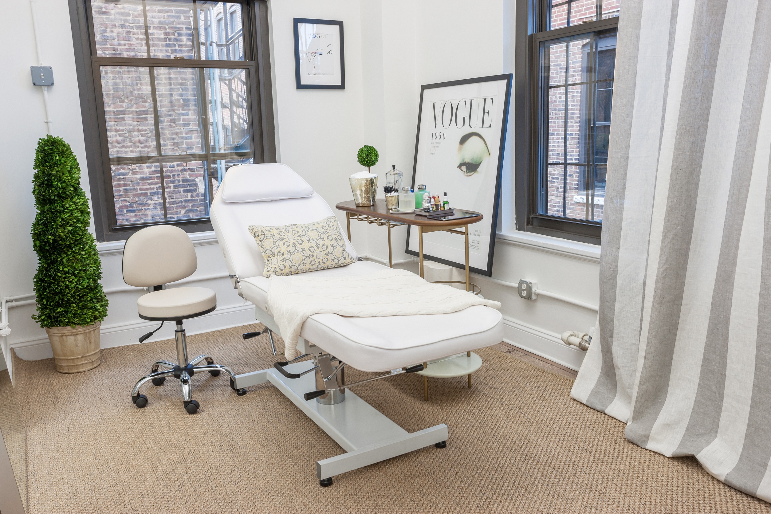 A treatment room with a reclining white chair, a stool, a side table with skincare products, framed art, and large windows with city views.