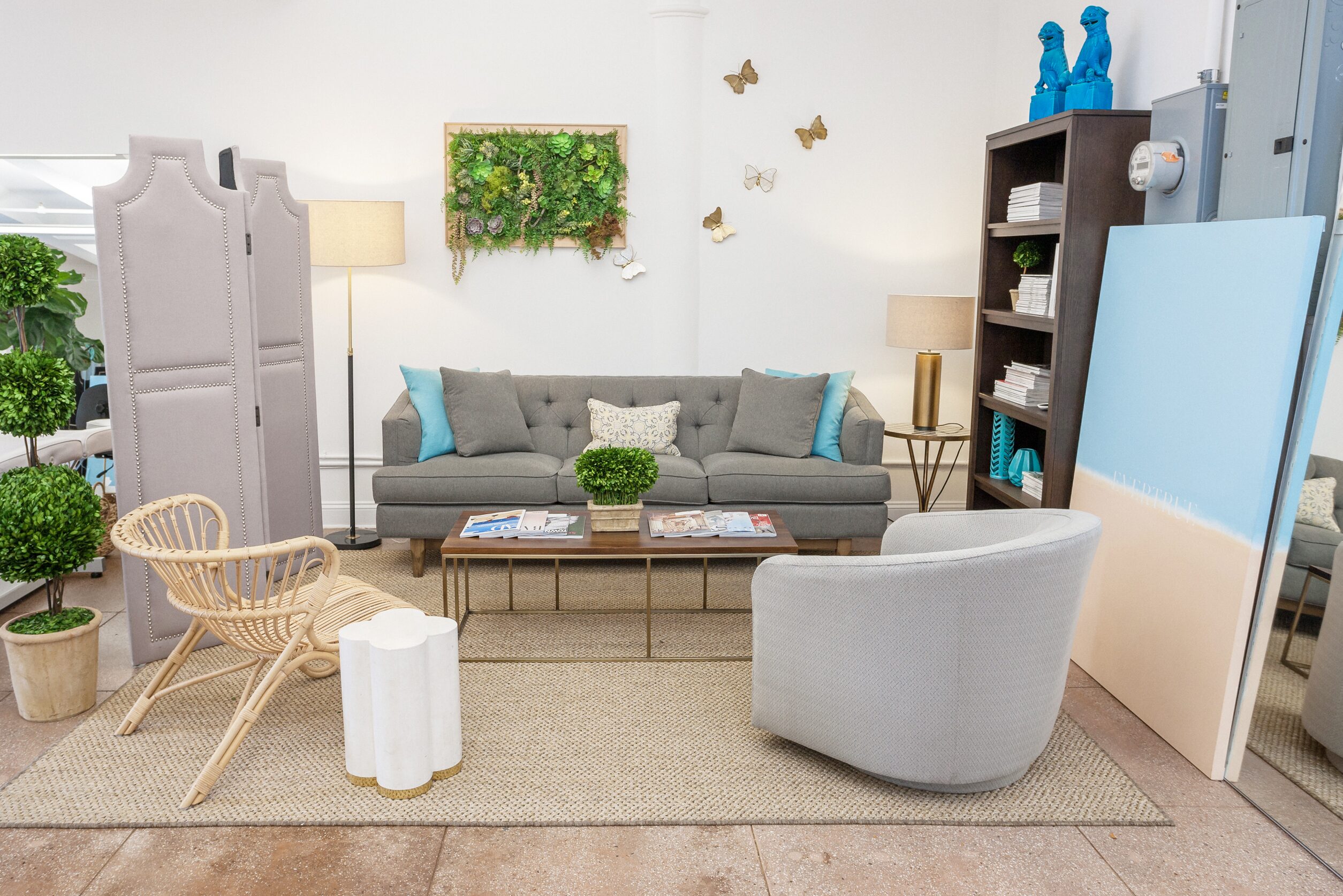 Modern living room with a gray sofa, light gray chair, rattan armchair, coffee table, bookshelf, decorative plants, and wall art.