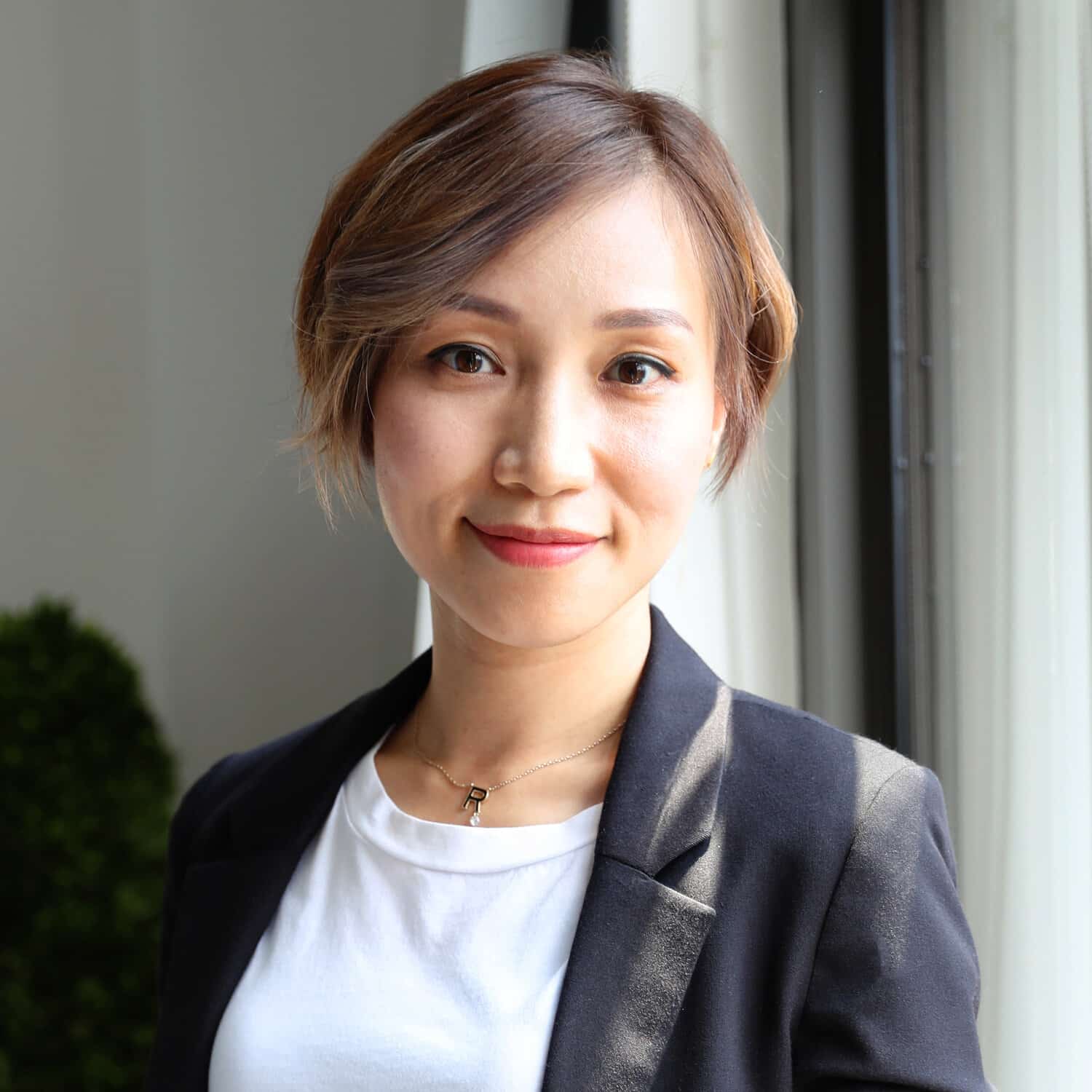 A woman with short brown hair wearing a black blazer and white shirt stands near a window, smiling slightly at the camera.