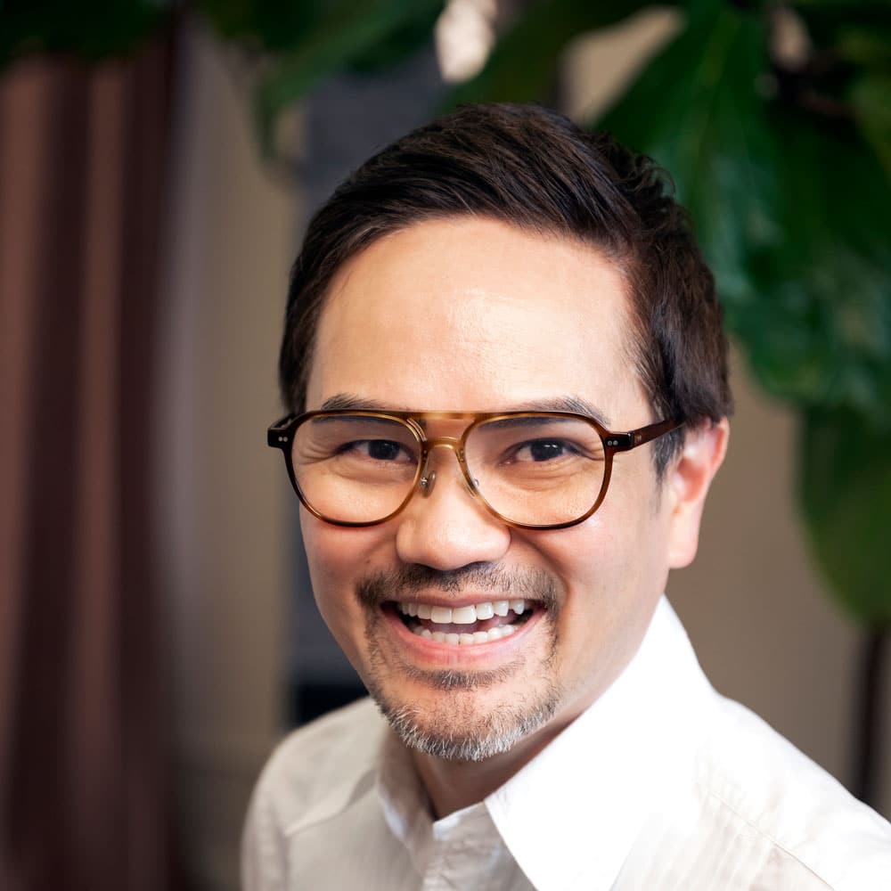 A man with short dark hair, glasses, and a goatee is smiling in a white collared shirt, with green leaves behind him—meet the expert beauty team at Evertrue Salon.