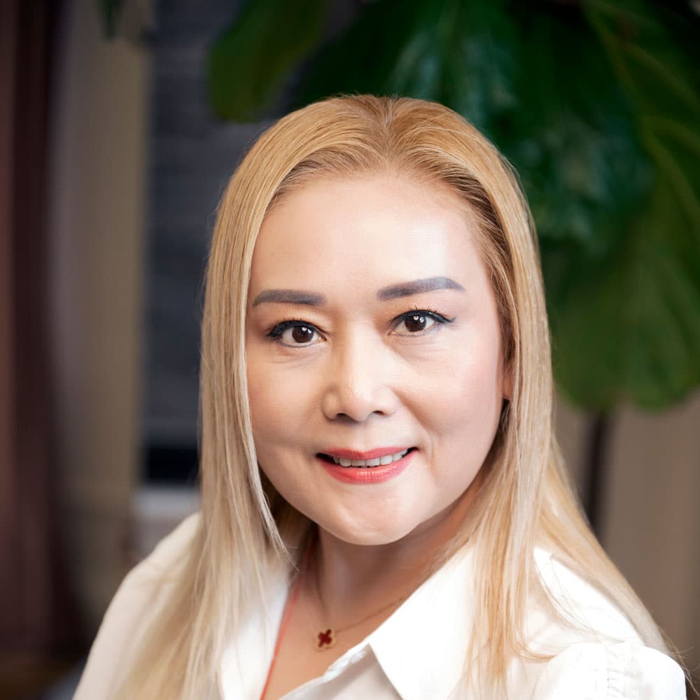 A woman with long blonde hair and a white shirt smiles at the camera, green leaves blurred behind her—Meet the Expert Beauty Team at Evertrue Salon.