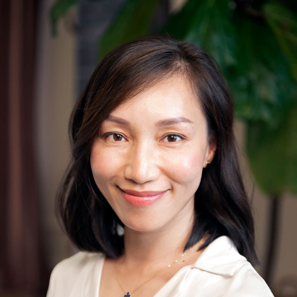 Meet the Expert Beauty Team at Evertrue Salon: A woman with shoulder-length dark hair and a white blouse smiles at the camera, with a large green plant in the background.