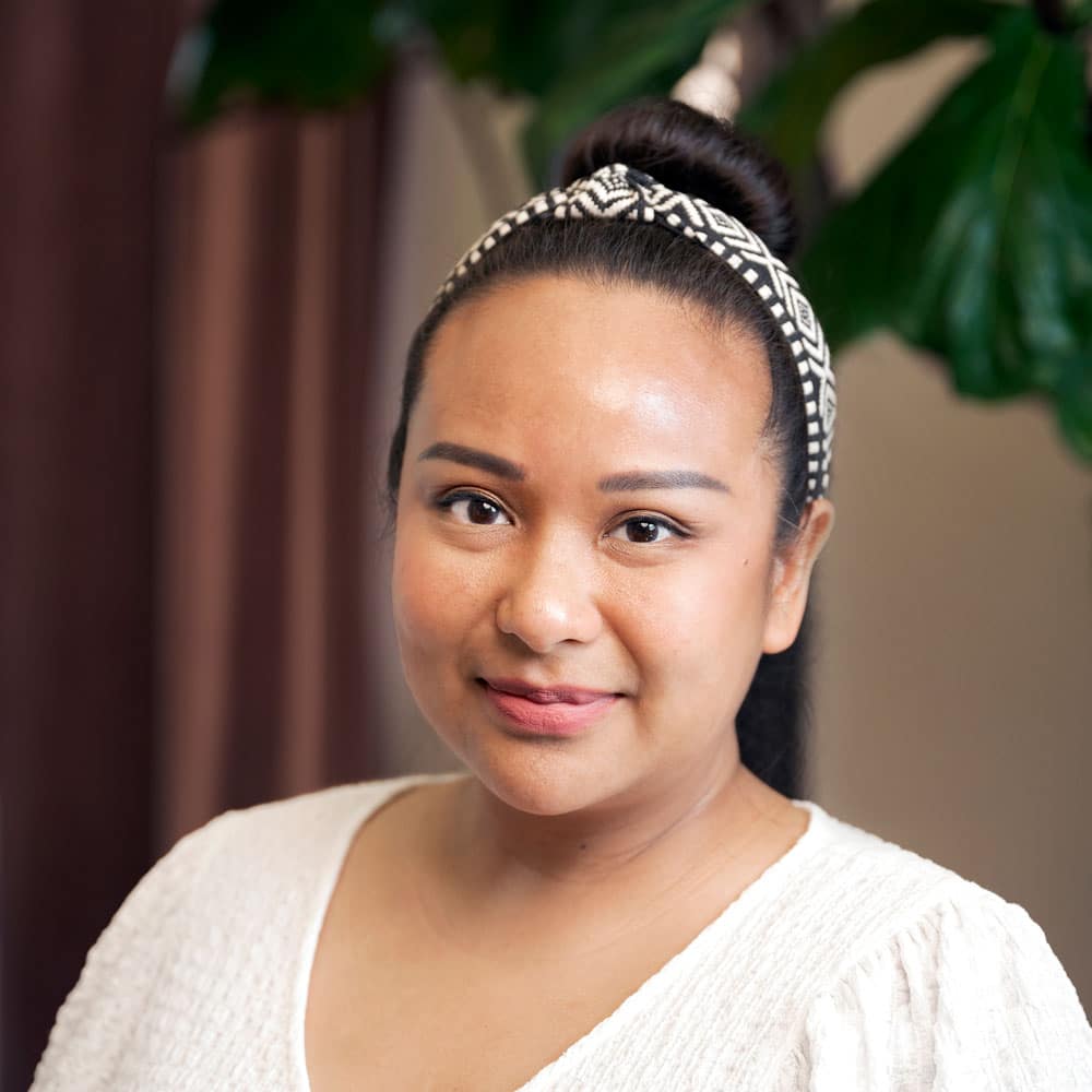 Meet the Expert Beauty Team at Evertrue Salon: A woman with a patterned headband and white blouse smiles slightly in front of a leafy green plant.