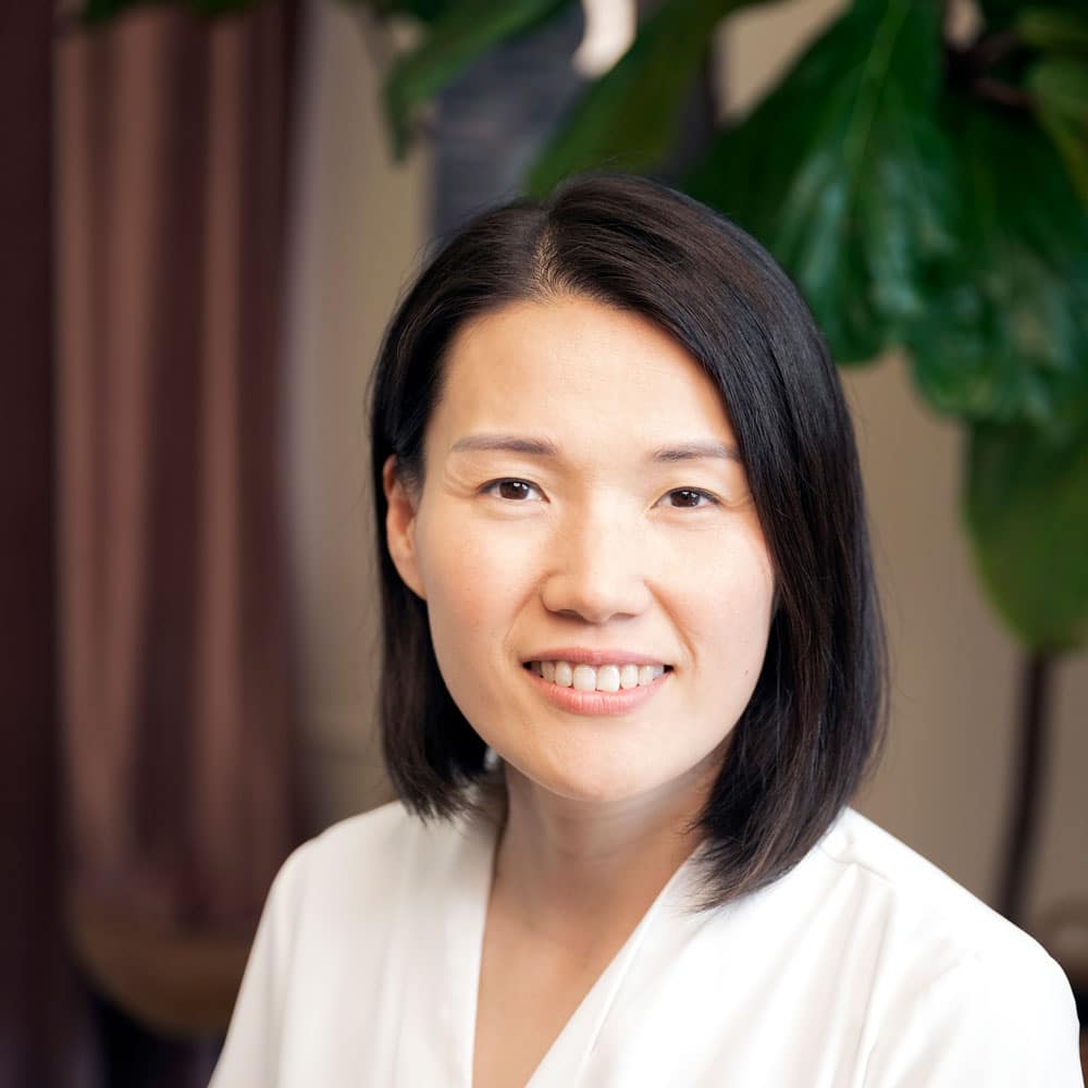 A woman with straight, shoulder-length black hair, wearing a white top, smiles at the camera. Green leaves and a curtain blur softly behind her—meet the expert beauty team at Evertrue Salon for your next transformation.