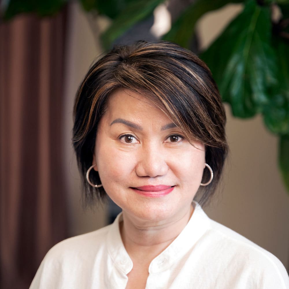 Meet the Expert Beauty Team at Evertrue Salon: Woman with short dark hair and hoop earrings, wearing a white top, smiling at the camera with large green leaves in the background.