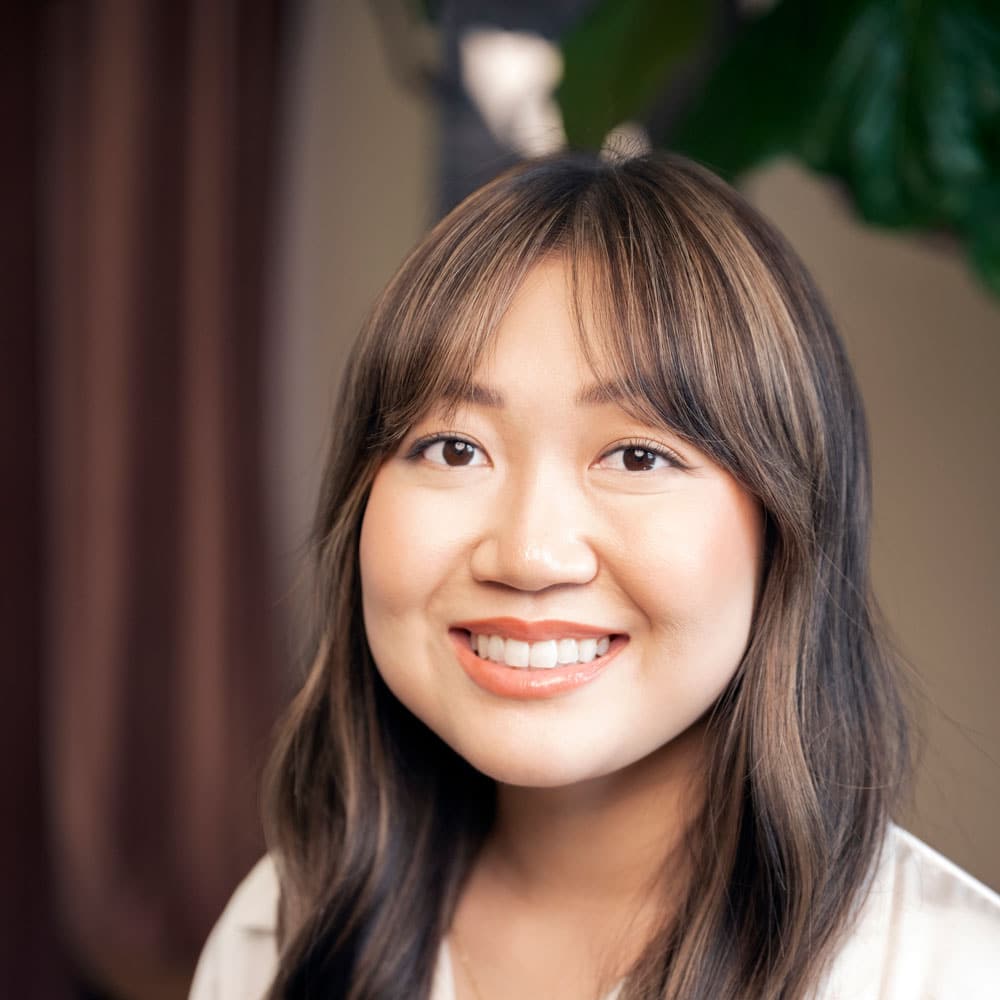 A young woman with long brown hair and bangs smiles at the camera, wearing a white top. A blurred background shows a plant and a curtain—meet the expert beauty team at Evertrue Salon for your next transformation.