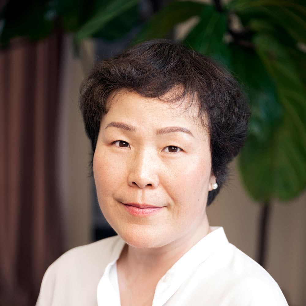 A woman with short, dark hair and a white blouse looks at the camera, standing indoors with green leaves in the background—an expert from the Meet the Expert Beauty Team at Evertrue Salon.
