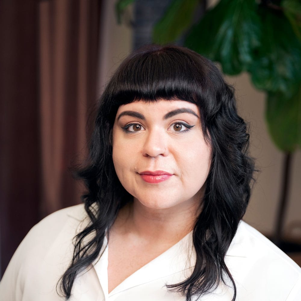 A person with black wavy hair and bangs, wearing a white top, poses indoors in front of a blurred background with a leafy green plant—just the kind of style perfected by the expert beauty team at Evertrue Salon.