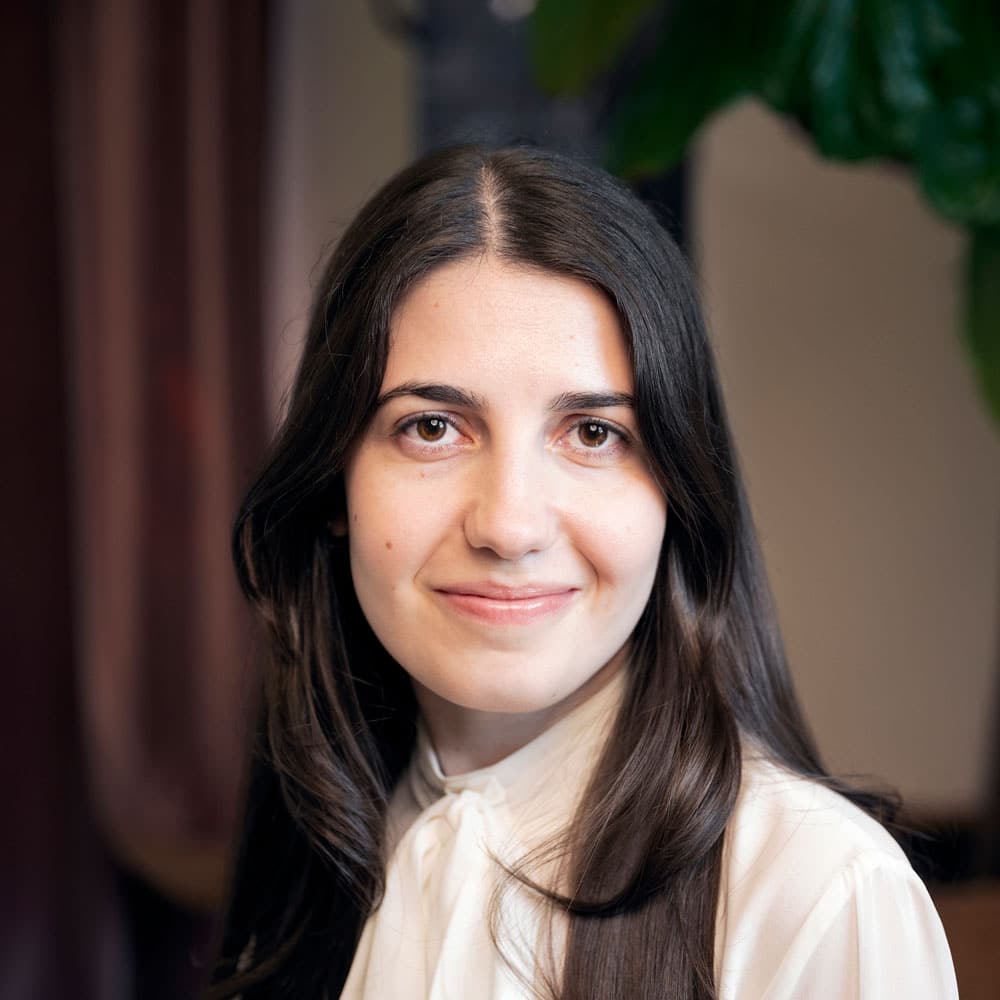 A woman with long dark hair and a white blouse smiles at the camera against a blurred indoor background, embodying the warmth of the Meet the Expert Beauty Team at Evertrue Salon.