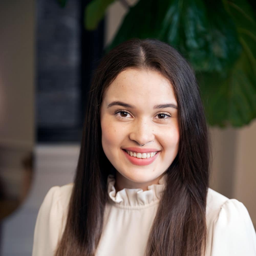 A young woman with long brown hair and a light blouse smiles at the camera, framed by a green leafy plant—meet the expert beauty team at Evertrue Salon.