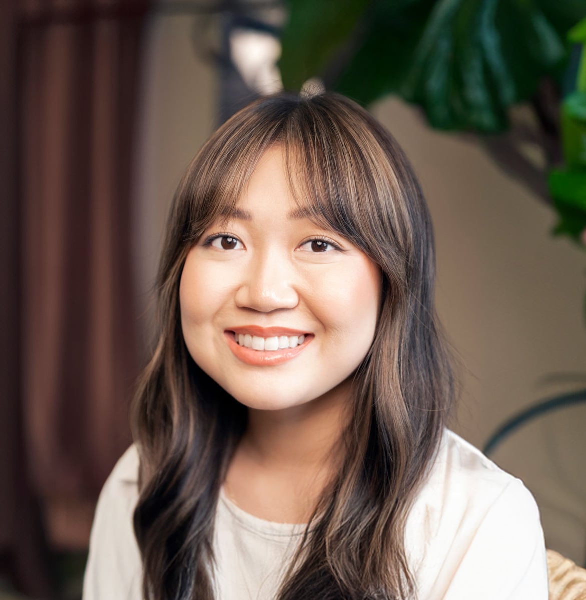 A woman with long, wavy brown hair and bangs smiles at the camera. She is wearing a light-colored top, surrounded by green leaves—a look perfected by the Meet the Expert Beauty Team at Evertrue Salon.