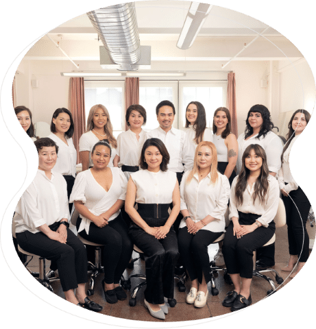 A group of fifteen people, mostly women, pose indoors wearing coordinated white tops and black pants or skirts, seated and standing in two rows—professionals specializing in microblading and nanobrows.