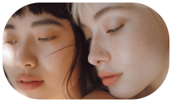 Two people with close faces look downward in soft lighting—one with dark hair and the other with light hair—creating a calm, intimate atmosphere that subtly highlights expertly shaped brows through microblading or nanobrows.