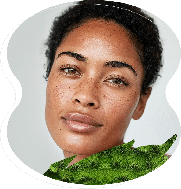 A woman with short curly hair and freckles looks at the camera, showing off expertly done nanobrows, with a green leafy plant partially in the foreground.