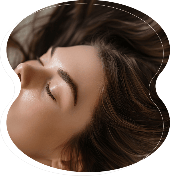 A close-up of a woman's face with closed eyes and smooth skin, framed by long brown hair, viewed through an abstract, rounded shape—her brows impeccably defined with nanobrows artistry.