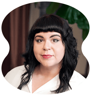 A person with wavy black hair, blunt bangs, and light makeup is wearing a white top and looking at the camera. A leafy green plant is in the background, capturing the fresh vibe of Evertrue salon Chicago.