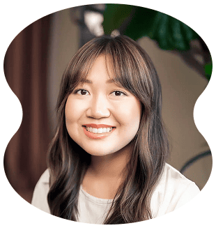 A woman with long brown hair and bangs, styled at Evertrue Salon, smiles at the camera in a white top, with a blurred indoor background.