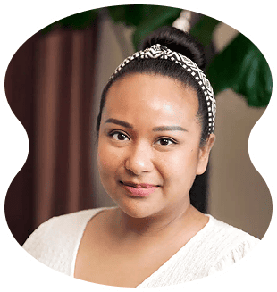 Woman with dark hair in a bun, wearing a patterned headband and white top, smiles slightly at evertrue salon, with a blurred indoor background and green plant visible.