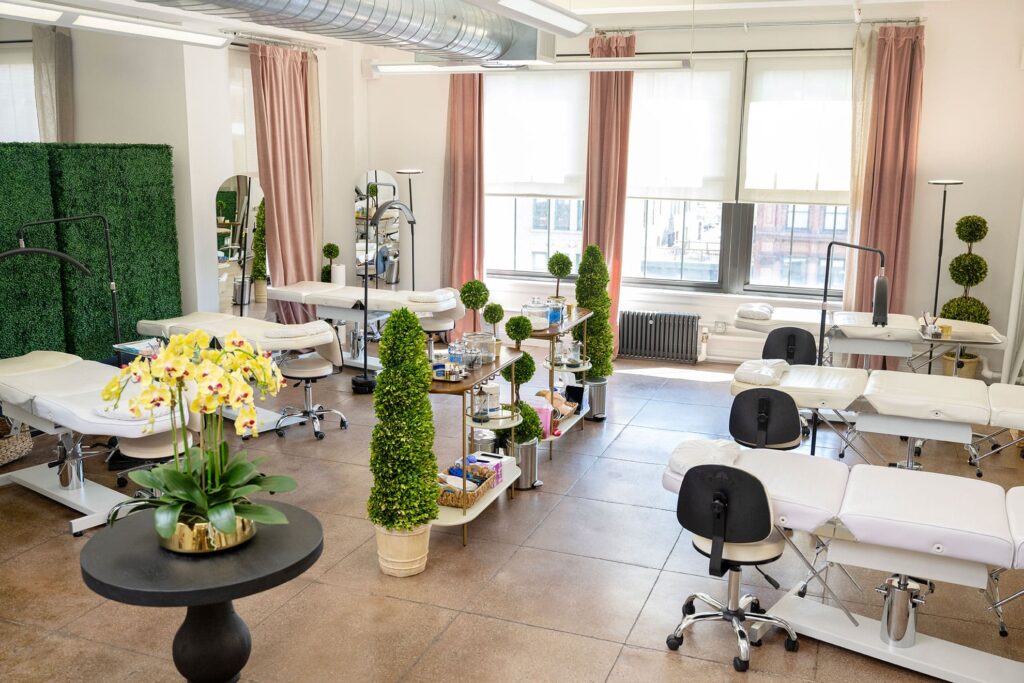 A bright, modern beauty treatment room with multiple reclined chairs, potted plants, supply trolleys, large windows, and pink curtains—perfect for sessions like those offered through the evertrue visiting artist program.