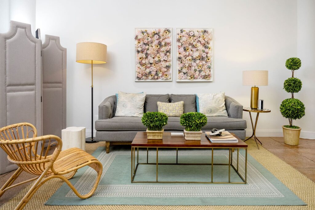 Modern living room with a gray sofa, floral wall art inspired by the Evertrue Visiting Artist Program, potted plants, a wicker chair, two floor lamps, and a wooden coffee table on a layered area rug.