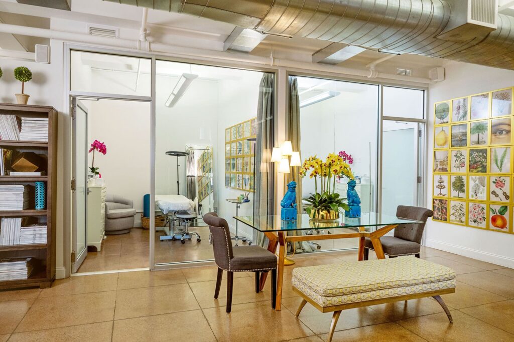 Modern office with a glass-walled room, a glass-top table featuring blue figurines, chairs, bench, bookshelves, and a wall decorated with botanical and facial art prints curated through the evertrue visiting artist program.