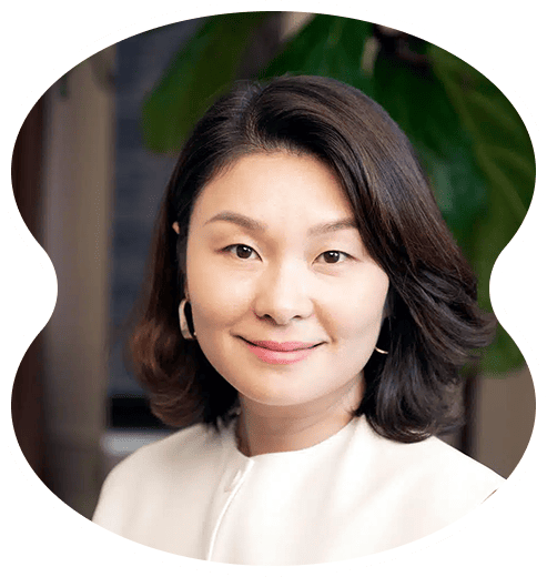 A woman with shoulder-length dark hair, wearing a white top and hoop earrings, smiles in front of a blurred background with a green plant—a welcoming image that reflects the inviting atmosphere about Evertrue Salon.