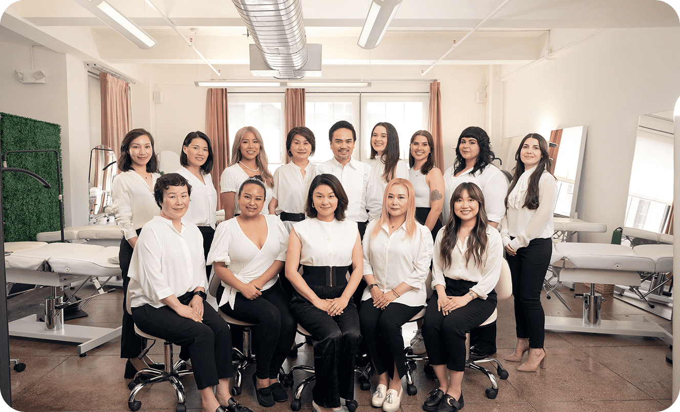 A group of fifteen people, mostly women, wearing white tops and black pants, pose together in a well-lit clinic or about Evertrue Salon setting with treatment beds in the background.