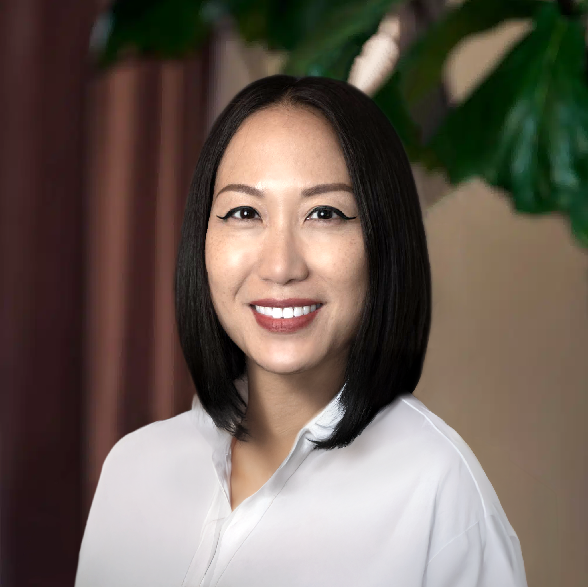 A woman with straight black hair and winged eyeliner smiles at the camera, wearing a white collared shirt. A large green plant is visible in the background, reflecting her interest in beauty care and after care tips.