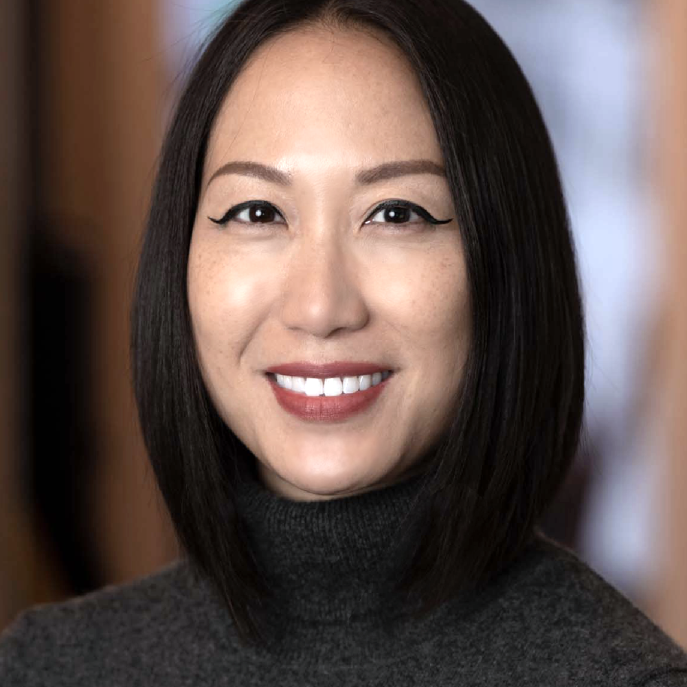 A woman with straight dark hair in a bob, wearing a dark turtleneck sweater, smiles at the camera against a blurred indoor background—meet the expert beauty team at Evertrue Salon.